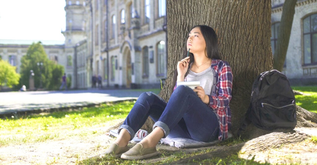 The image shows an international student thinking about GMAT exam outside fo the university building